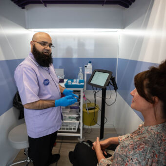 Male NHS-Galleri trial nurse in a cubicle on a trial unit, explaining the blood sampling process to a female participant
