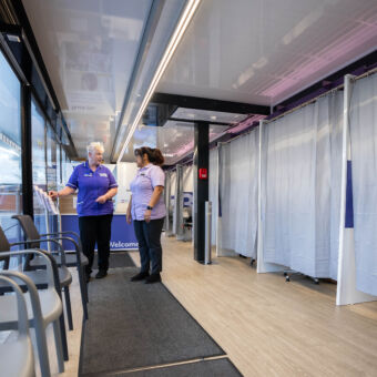 Two nurses talk in the waiting room of an NHS-Galleri trial mobile unit, with cubicles for appointments behind them